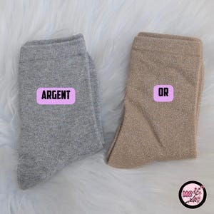 May include: Two pairs of shimmering socks, one silver and one gold, are displayed on a fluffy white surface. The silver sock has a pink label that says "ARGENT", and the gold sock has a pink label that says "OR".