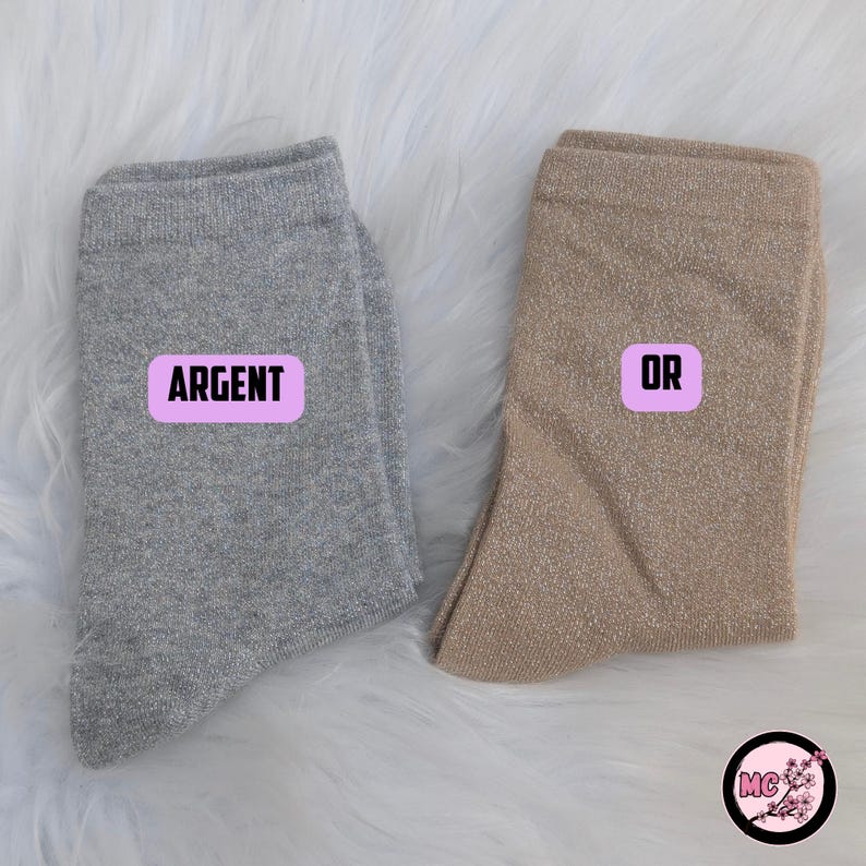 May include: Two pairs of shimmering socks, one silver and one gold, are displayed on a fluffy white surface. The silver sock has a pink label that says "ARGENT", and the gold sock has a pink label that says "OR".