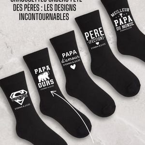 Peut inclure: Chaussettes noires avec des textes et des graphiques blancs. Les chaussettes comportent des phrases comme "Super Papa", "Papa Ours" et "Meilleur Papa du Monde". Le texte "D'Emma & Jules" apparaît sur chaque chaussette. Le texte en français est également présent.