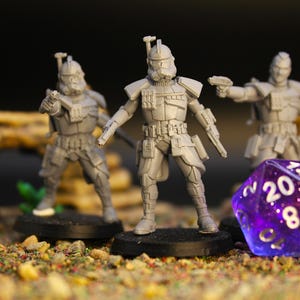 Op de afbeelding: Drie grijze miniatuurfiguren van soldaten met wapens, staand op zwarte bases, staan voor een paarse, veelzijdige dobbelsteen. De figuren zijn gedetailleerd met pantser en helmen. De sc&egrave;ne speelt zich af op een getextureerd oppervlak met kleine stenen en vegetatie.