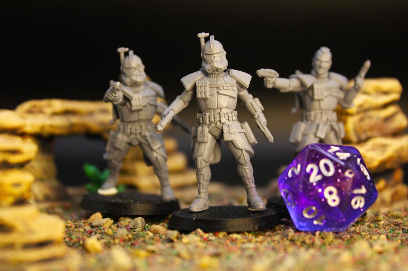 Op de afbeelding: Drie grijze miniatuurfiguren van soldaten met wapens, staand op zwarte bases, staan voor een paarse, veelzijdige dobbelsteen. De figuren zijn gedetailleerd met pantser en helmen. De sc&egrave;ne speelt zich af op een getextureerd oppervlak met kleine stenen en vegetatie.