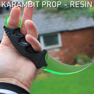 Op de afbeelding: Zwart en groen karambit-rekwisiet van hars. Het gebogen lemmet is doorschijnend groen en het handvat is zwart. De tekst "KARAMBIT PROP - RESIN" staat bovenaan de afbeelding.