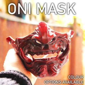 May include: Red Oni mask with a fierce expression, featuring sharp teeth and detailed carvings. The mask is held in a hand, with the text "COLOUR OPTIONS AVAILABLE" visible below.