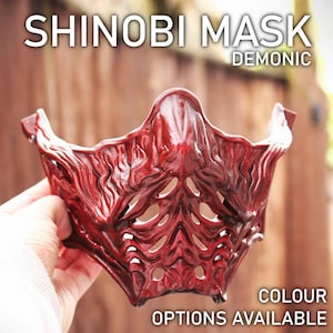 Può includere: Una maschera shinobi rossa in stile demoniaco con un design testurizzato dettagliato. La maschera ha una grande bocca aperta e una serie di piccoli fori aperti intorno agli occhi e al naso. Il testo "SHINOBI MASK DEMONC" è visibile nella parte superiore dell'immagine.