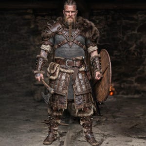 May include: A full-body shot of a person in a Viking warrior costume. The costume includes brown leather armor, a fur collar, and a chainmail shirt. The warrior holds an axe in one hand and a wooden shield in the other.