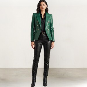 Könnte beinhalten: Ein grüner Blazer mit Krokodiltextur und goldenen Knöpfen, kombiniert mit einer schwarzen Hose und Stiefeln. Das Outfit umfasst ein schwarzes Hemd und einen Gürtel. Der zweireihige Blazer ist ein zentrales Element.