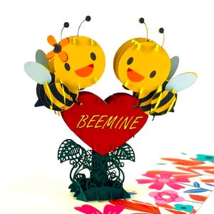 Könnte beinhalten: Eine Pop-up-Grußkarte mit zwei Cartoon-Bienen, die ein rotes Herz mit dem Text "BEEMINE" halten. Die Bienen sind gelb und schwarz mit lächelnden Gesichtern. Die Karte hat eine grüne Basis mit floralen Akzenten.