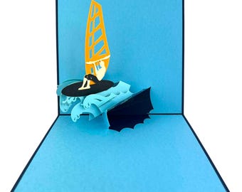 May include: A pop-up card featuring a windsurfer on blue waves with an orange sail. The card is blue with a black border, creating a 3D effect. The windsurfer is black and white.