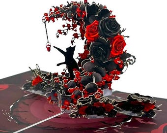 May include: A pop-up card featuring a black cat reaching for a hanging red gem. The card has a crescent-shaped arrangement of black and red roses and foliage. The card is open, revealing a layered design with a dark red and black color scheme.