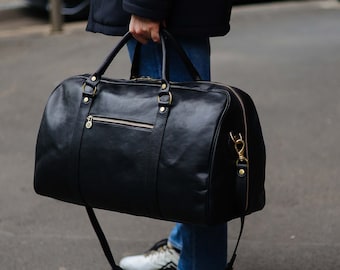 Può includere: Un borsone da viaggio in pelle nera con finiture in metallo dorato, inclusa una tasca frontale con cerniera, anelli laterali di fissaggio e una tracolla rimovibile. La borsa presenta due manici superiori e una finitura liscia con cuciture.