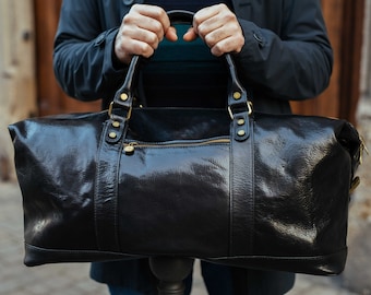 Op de afbeelding: Een glanzende zwarte leren duffeltas met goudkleurige hardware, inclusief ritsen en klinknagels. De tas heeft twee handvatten aan de bovenkant en een schouderriem. De tas wordt door een persoon vastgehouden.
