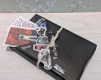 May include: A black paper envelope tied with twine, featuring a collection of space-themed stickers and postcards. The stickers include images of a rocket, astronaut, and space jars. The envelope has the words "Vintage Sci-fi" and "Space" printed on it.