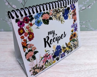 May include: A white recipe book with a black spiral binding and the words "my Recipes" in black script. The book is decorated with a floral border of colorful flowers, including pink, blue, and yellow blooms.