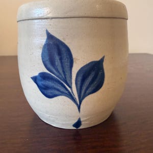 May include: A cylindrical stoneware crock with a speckled off-white finish and a smooth, rounded rim. The exterior features a hand-painted cobalt blue design of three pointed leaves arranged in a cluster, with a small detached blue droplet below.