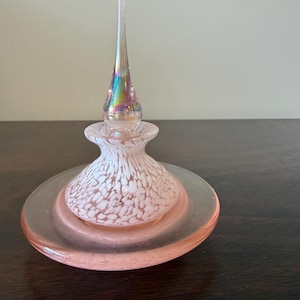 May include: A glass perfume bottle featuring a wide, flat, translucent pink base and a rounded body with a white speckled pattern. The bottle is topped with a tall, slender, clear glass stopper that displays an iridescent, rainbow-like sheen.
