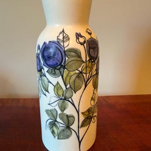 May include: A cylindrical ceramic vase with a cream-colored base featuring a hand-painted botanical illustration of purple rose blossoms and green leaves with dark outlines. The vase has a narrow neck and a wide, flat base for stability.
