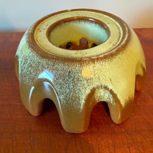 May include: A ceramic candle warmer featuring a dome-shaped body with a scalloped base and a central circular opening. The exterior has a glossy glaze with a gradient transition from a deep brown at the bottom to a light yellow-green at the top.