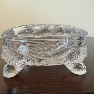 May include: A clear glass trinket box with a rectangular shape and a removable lid. The exterior features a molded, swirling wave-like pattern throughout the body and lid. The box is supported by four distinct, textured feet at each corner.