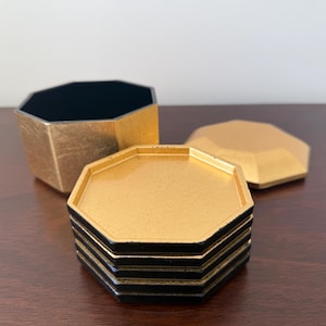 May include: A set of gold and black octagonal coasters and a matching container. The coasters are stacked, and the container is open, revealing a black interior. A gold lid is also present.