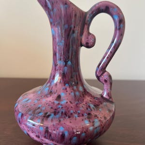 May include: A small purple and blue ceramic pitcher with a speckled glaze. The pitcher has a rounded body and a curved handle.