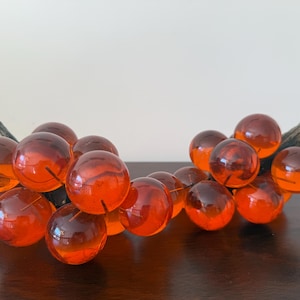 May include: A decorative cluster of orange-red glass grapes with a dark brown stem. The glass spheres are arranged to resemble a bunch of grapes, perfect for home decor.