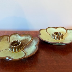 May include: Two ceramic candle holders shaped like flowers with a central cylindrical socket. The petals feature a light green glaze with brown edges and a brown center. The finish is glossy, and the edges of the petals are scalloped and rounded.