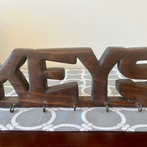 May include: Wooden key holder with the word "KEYS" carved into it. The holder has five hooks for hanging keys.