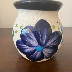 May include: A ceramic vase with a speckled white base and a dark blue rim. The vase features a large, stylized blue flower with a turquoise center. Green leaf accents are scattered around the flower, creating a decorative floral design.