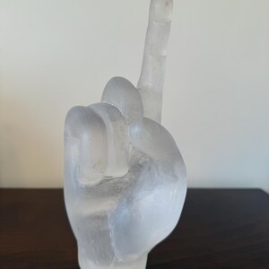 May include: A clear, frosted glass sculpture of a hand with the index finger pointing upwards. The sculpture is set against a neutral background and rests on a dark wooden surface. The hand is detailed, with visible knuckles and a rounded base.