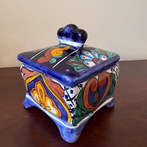 May include: A square, ceramic trinket box with a lid, featuring a vibrant, hand-painted design. The box is primarily blue, with colorful floral and geometric patterns in red, yellow, and green. It has a small, cloud-shaped handle on the lid and sits on four small feet.