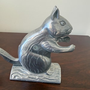 May include: A silver-colored squirrel nutcracker. The squirrel is perched on a log-shaped base, with its arms extended. The tail curves upward. The nutcracker is made of metal and has detailed fur and facial features.