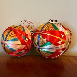 May include: Two spherical temari balls wrapped in vibrant red, gold, blue, green, and white silk thread. Each ball features intricate geometric patterns and a small tag that reads Made in Japan, with red hanging strings attached to the top.