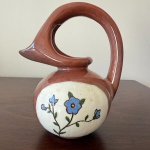 May include: A ceramic pitcher with a distinctive handle and a bulbous body. The handle and top are a reddish-brown color, while the body is white with blue floral designs. The pitcher is likely used for decorative purposes.