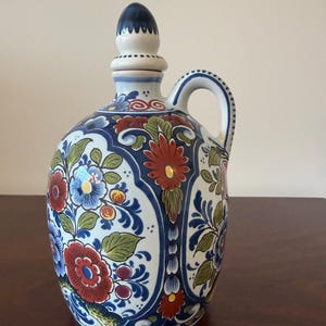May include: A ceramic pitcher with a handle and a stopper. The pitcher is white with a blue floral design and red, blue, and yellow flowers. The stopper is dark blue and white.