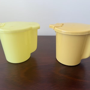 May include: Two yellow plastic containers with lids. One is a light yellow, the other is a darker yellow. Each container has a curved handle-like extension on the side and a hinged lid with a small tab.
