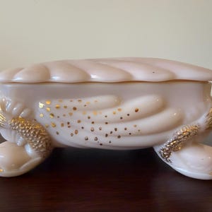 May include: A pale pink, lidded trinket box with a seashell design. The box has a textured surface with gold accents and rests on three rounded feet. Gold flecks decorate the side of the box.