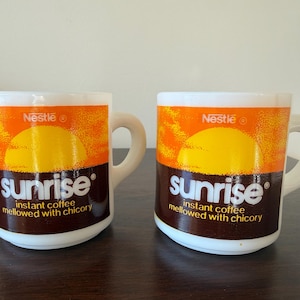 May include: Two white ceramic coffee mugs with the "Sunrise" logo. The mugs feature a yellow sun graphic against an orange background, with the Nestle logo and text "instant coffee mellowed with chicory".