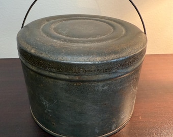 May include: A vintage, round metal container with a lid and a wire handle. The container has a dark, weathered appearance, showing signs of age and use. The handle arches over the top, and the lid fits snugly. The overall aesthetic suggests a rustic or antique style.
