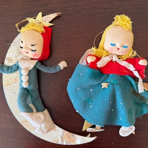 May include: Two vintage-style ornaments featuring dolls with yellow yarn hair. One doll wears a teal suit and rests on a crescent moon. The other doll wears a red top and teal skirt, positioned against a star. Both have painted faces and felt fabric.
