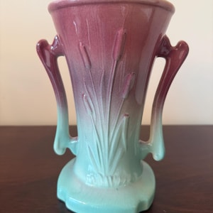 May include: A ceramic vase featuring a two-tone gradient design that transitions from a deep magenta at the top to a light mint green at the base. The vase has two side handles and a molded relief pattern of cattails on the front surface.
