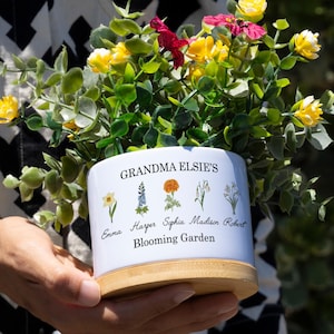 May include: White ceramic planter with a wooden base. The planter has a floral design with the text "Grandma Elsie's Blooming Garden" and the names "Emma, Harper, Sophia, Madison, Robert" printed on it. The planter is filled with a variety of yellow and red flowers.
