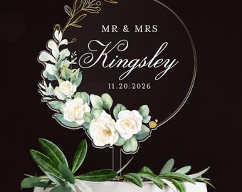 Könnte beinhalten: Ein runder Tortenstecker aus klarem Acryl mit einem aufgedruckten Blumenkranz aus weißen Rosen und grünen Blättern. Der Text lautet MR & MRS Kingsley 11.20.2026 in weißer und goldener Schrift. Der Stecker steckt in einer weiß glasierten Torte.