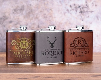 May include: Three personalized stainless steel hip flasks with leather coverings. The flasks are engraved with the names Michael, Robert, and Richard, along with their initials and the date 11.04.2023. The flasks are brown, gray, and brown.