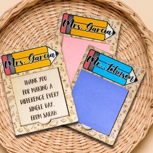 May include: Two wooden pencil-shaped note holders with a space for a sticky note. The top of the pencil is decorated with a yellow and blue pencil and the text "Mrs. Garcia" and "Mr. Johnson". The bottom of the pencil has a brown background with a variety of school-themed icons. The note holder on the left has a pink sticky note with the text "THANK YOU FOR MAKING A DIFFERENCE EVERY SINGLE DAY. FROM SARAH". The note holder on the right has a blue sticky note.