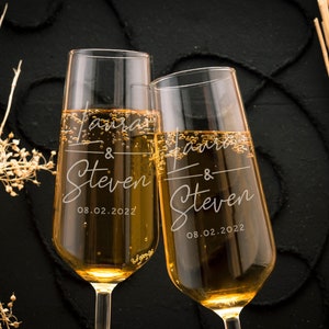 May include: Two champagne flutes filled with a golden liquid, likely champagne or sparkling wine. Each glass is engraved with the names "Laura & Steven" and the date "08.02.2022". The glasses are set against a dark background with decorative elements.