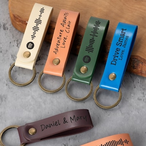 May include: Five leather keychains with different colors and personalized messages. The keychains feature a metal ring and a bar with a Spotify code and a personalized message. The messages include "Adventure Awaits Love, Clair", "Drive Smart Love, James", "Daniel & Mary", and two other messages with Spotify codes.