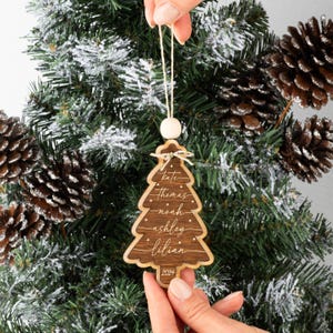 May include: A dark brown wooden Christmas tree ornament with a light wood border. The ornament features the names Kate, Thomas, Noah, Ashley, and Lilian engraved in white script, with small star accents and the year 2024 at the base.