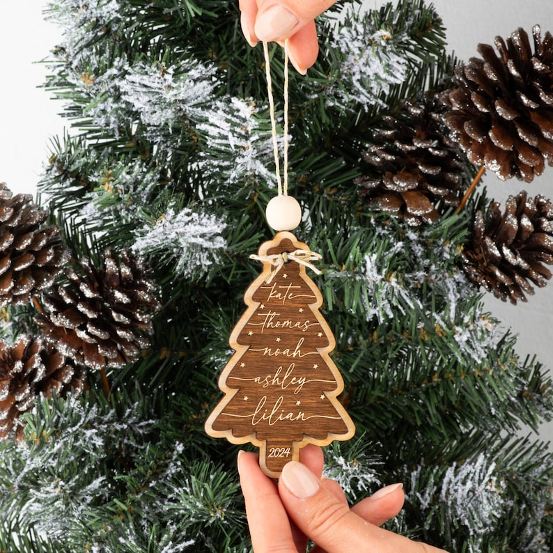 May include: A dark brown wooden Christmas tree ornament with a light wood border. The ornament features the names Kate, Thomas, Noah, Ashley, and Lilian engraved in white script, with small star accents and the year 2024 at the base.