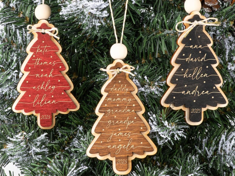 May include: Three wooden Christmas tree ornaments in red, brown, and black. Each ornament features a layered design, a white wooden bead, a twine string, and a small bow. The ornaments are engraved with names and the year 2025 in white script.