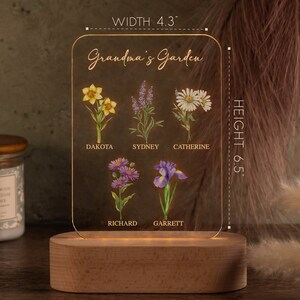 May include: A personalized acrylic garden sign with a wooden base. The sign features the text "Grandma's Garden" and illustrations of various flowers, with names like Dakota, Sydney, and Catherine. The sign is 4.3 inches wide and 6.5 inches tall.